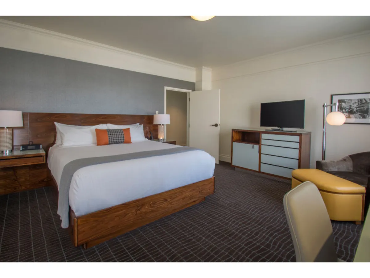 This image showcases a stylish, modern hotel room in downtown Portland featuring a plush bed with crisp linens, a sleek nightstand, a flat-screen television across from the bed, and a cozy armchair by the window. The space feels inviting, perfect for unwinding after a day in the city, whether guests are sipping morning coffee in bed, reading in the armchair, or enjoying a movie on a rainy night.