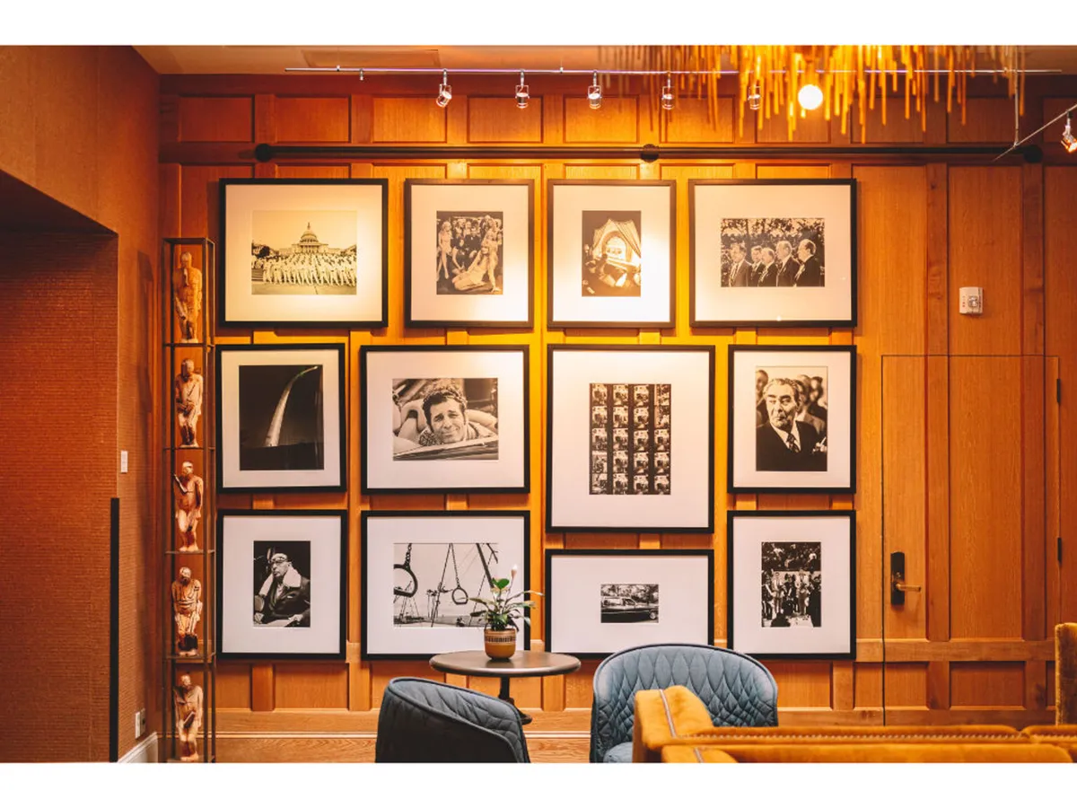 This image showcases a gallery wall filled with framed photographs, a small round table, blue armchairs, and rich wooden paneling. The photographic art is the focal point, signaling the curated and layered artistic sensibility highlighted in the section. The overall mood is nostalgic and sophisticated, inviting guests to engage with the artwork as soon as they arrive.