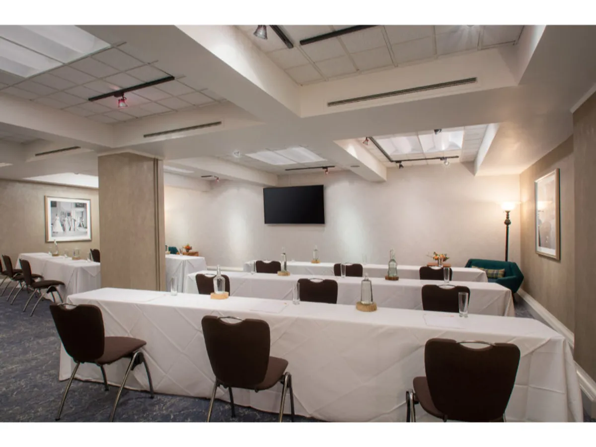 A well-lit meeting space featuring long tables arranged for collaboration, a wall-mounted television for presentations, and a stylish lamp in the corner, providing a focused yet comfortable environment for business travelers.