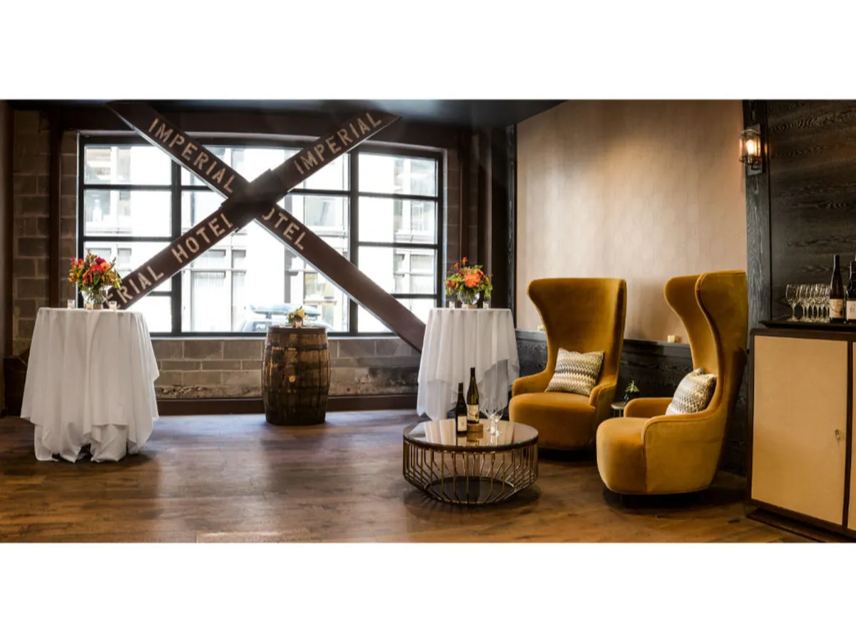Two stylish armchairs face small round tables adorned with floral arrangements. A barrel adds a touch of rustic charm, while a large window reveals the 'IMPERIAL HOTEL' sign outside, providing a direct visual connection between the cozy interior and the urban environment beyond.