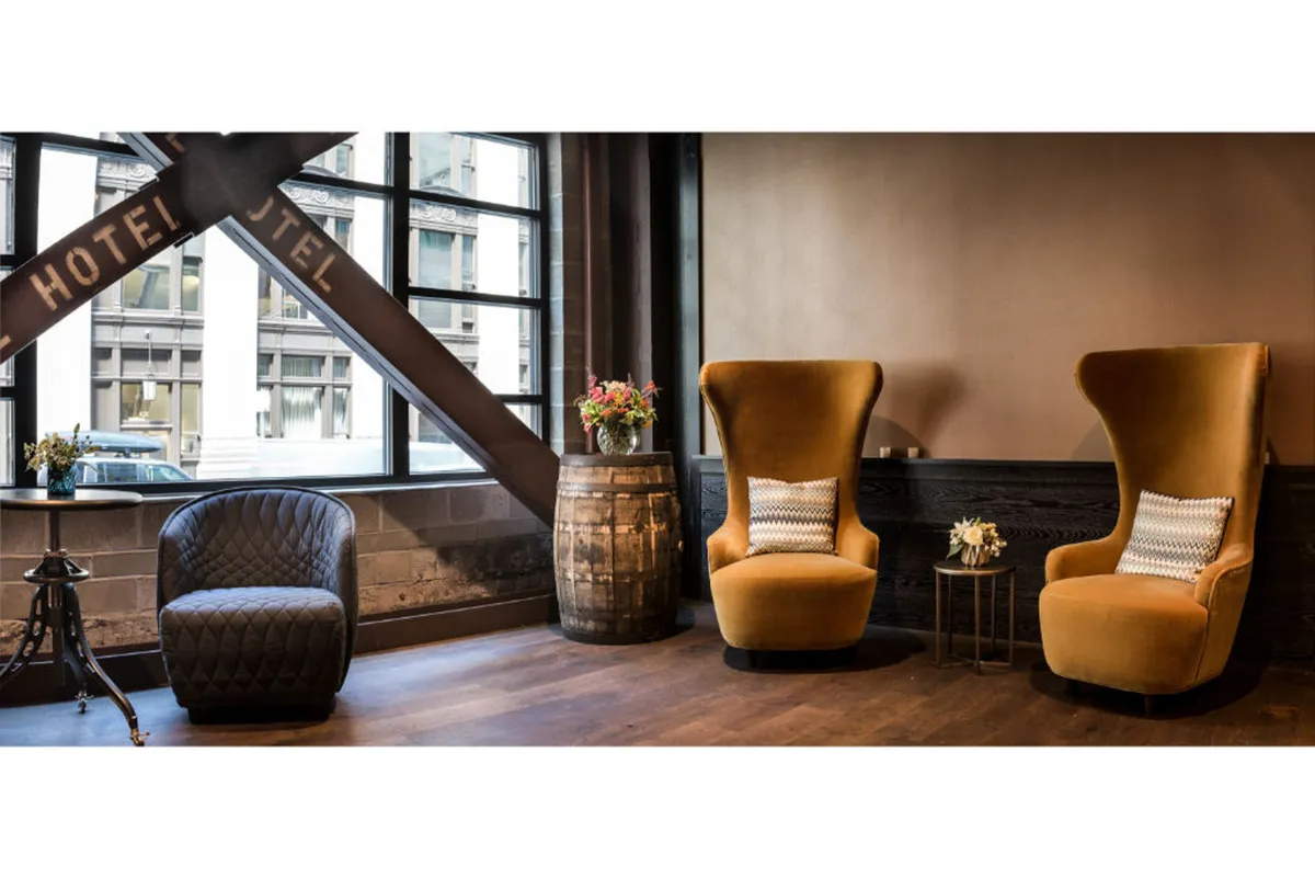 Large velvet chairs surround a wooden barrel repurposed as a table, with decorative flowers and a stylish side table completing the setting. The room is designed for comfort and reflection, with glimpses of the city through the window.