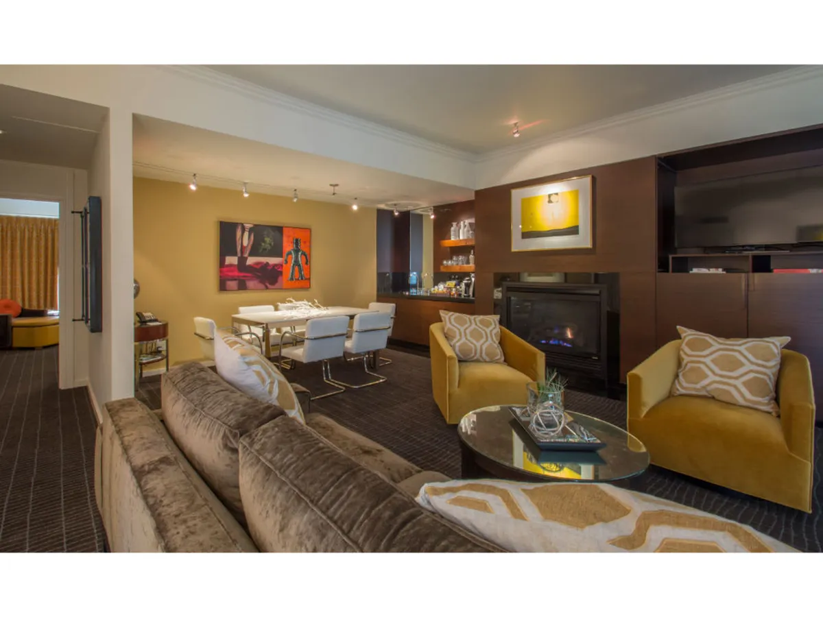 A spacious, well-lit living room featuring a modern sofa, an elegant dining table, prominent wall art, and a cozy gas fireplace. Large windows bring in ample natural light, creating an inviting atmosphere ideal for socializing or relaxing.