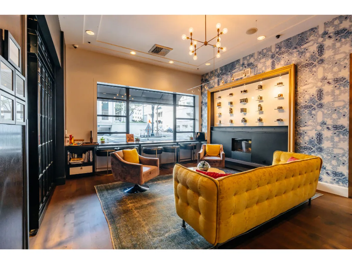 A bright, contemporary seating area with a yellow tufted sofa, wooden coffee table, decorative wall with framed items, and expansive windows that fill the space with natural light. The area is designed for both relaxation and work, offering a blend of comfort and style.