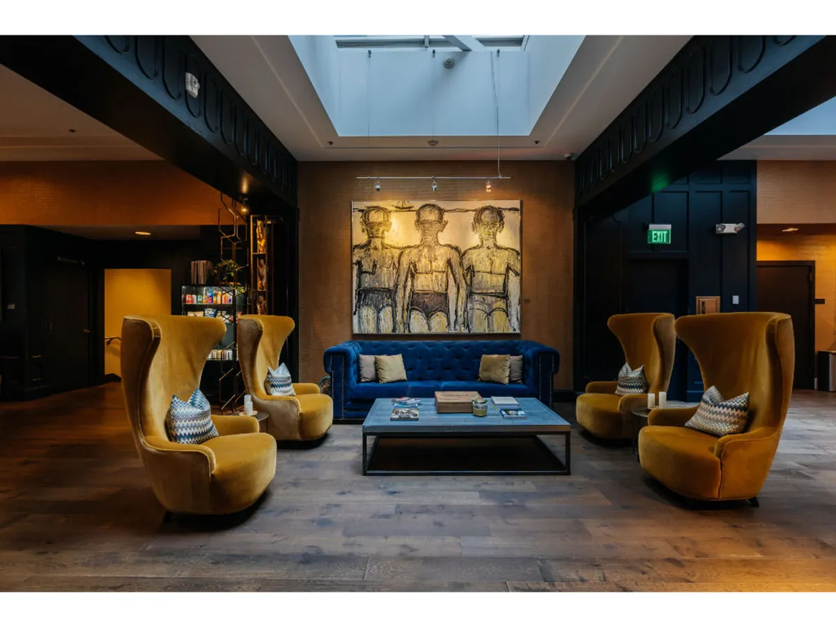 A cozy lounge featuring a plush blue velvet sofa, cheerful yellow armchairs, and art-adorned walls, centered around a coffee table. The scene feels comfortable, stylish, and inviting—ideal for relaxing with your pet after an active outing.