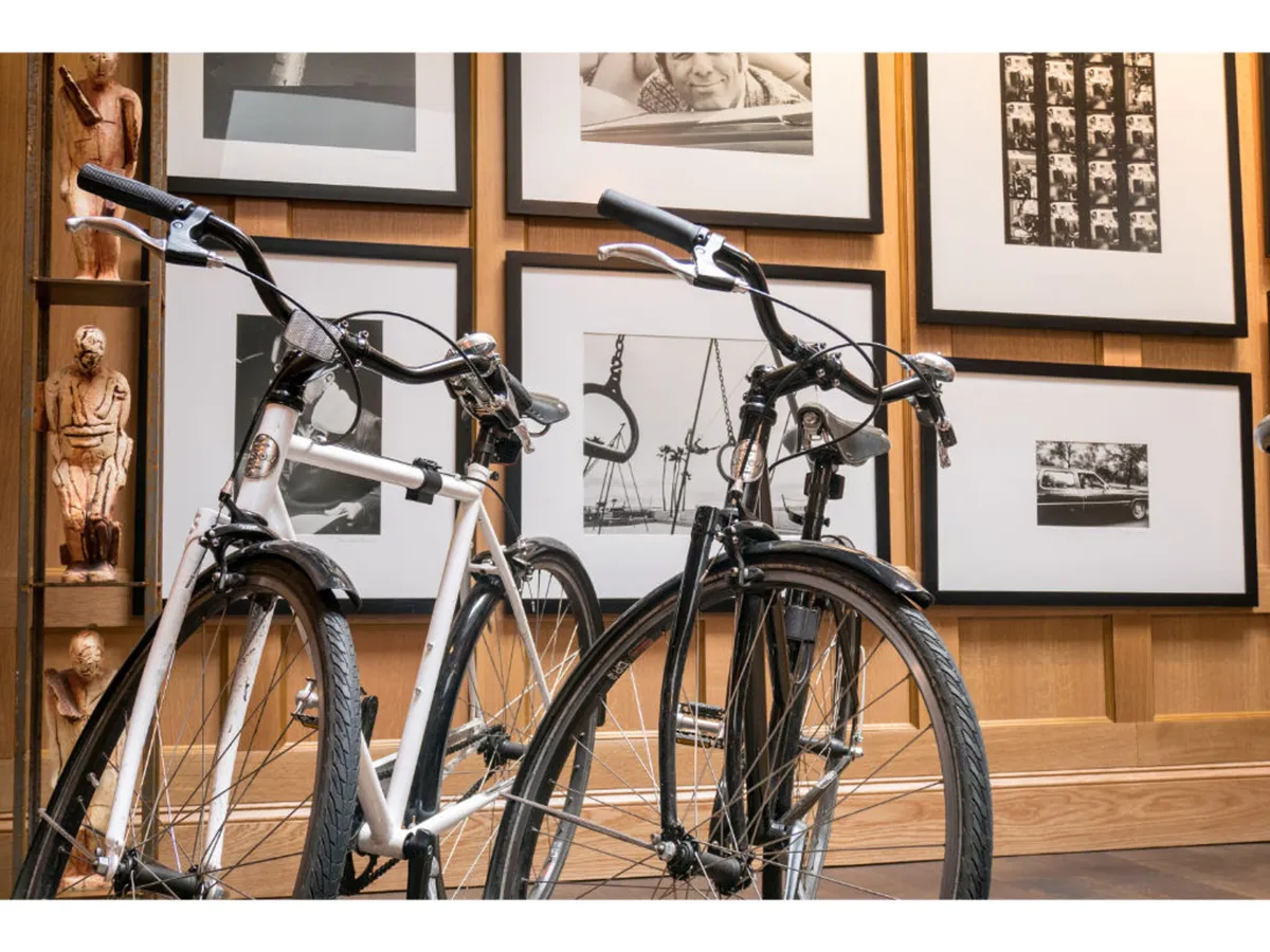 This image features two stylish bicycles (one white, one black) set against a backdrop of photographic artwork and warm wooden textures. The scene exudes a creative and contemporary vibe, suggesting active exploration and the hotel’s connection to local culture and amenities.