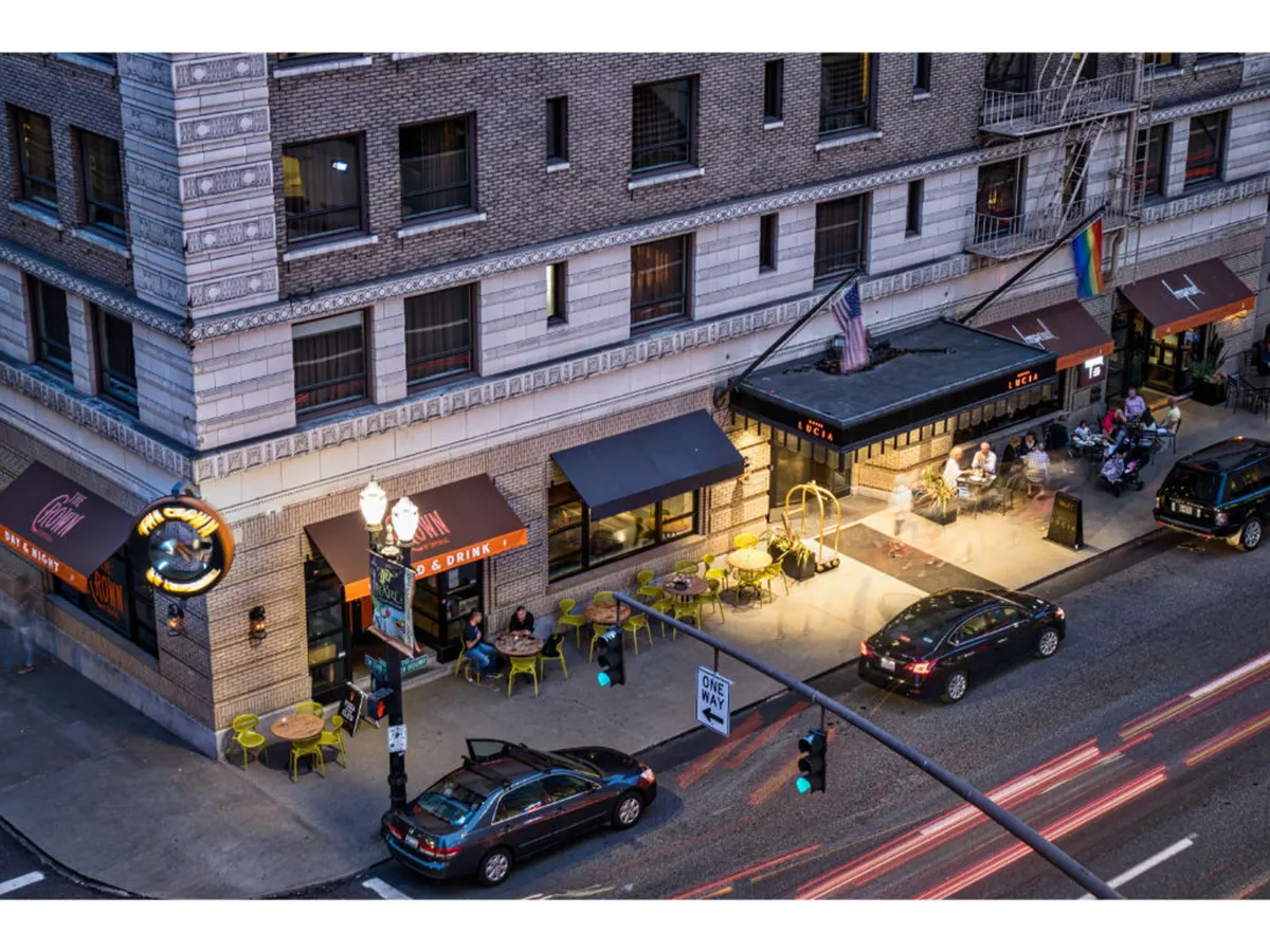 A lively exterior shot featuring an outdoor seating area, restaurant signage, and visible street traffic. The atmosphere is vibrant, suggesting accessibility to the surrounding urban environment and opportunities for socializing and people-watching.