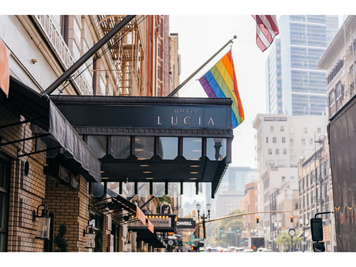 This exterior shot captures the Hotel Lucia sign prominently displayed above the entrance, with a rainbow flag and American flag adding vibrancy. The building's architectural details are visible, and the bustling downtown atmosphere is palpable, hinting at immediate access to Portland’s urban culture.