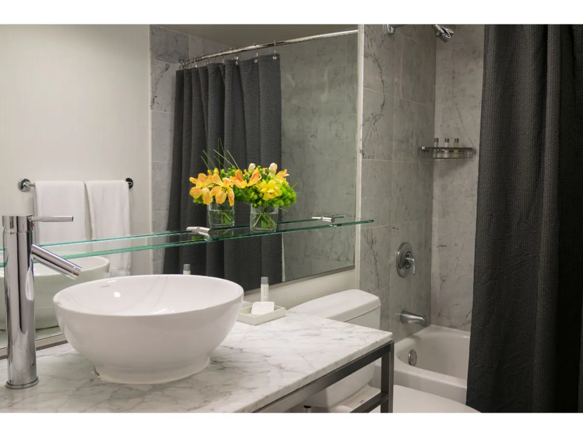 A serene, modern bathroom featuring a sleek sink and marble countertop, with a delicate flower arrangement and a glass shelf. The ambiance suggests a peaceful space suitable for self-care routines and relaxing baths.
