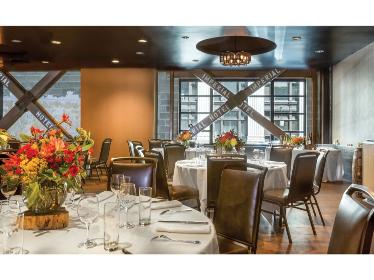 A refined dining area with round tables adorned with floral arrangements, basking in natural light from large windows that offer a sweeping city view. Elegant chandeliers hang overhead, and the space feels welcoming and open—ideal for mingling or relaxing after urban adventures.
